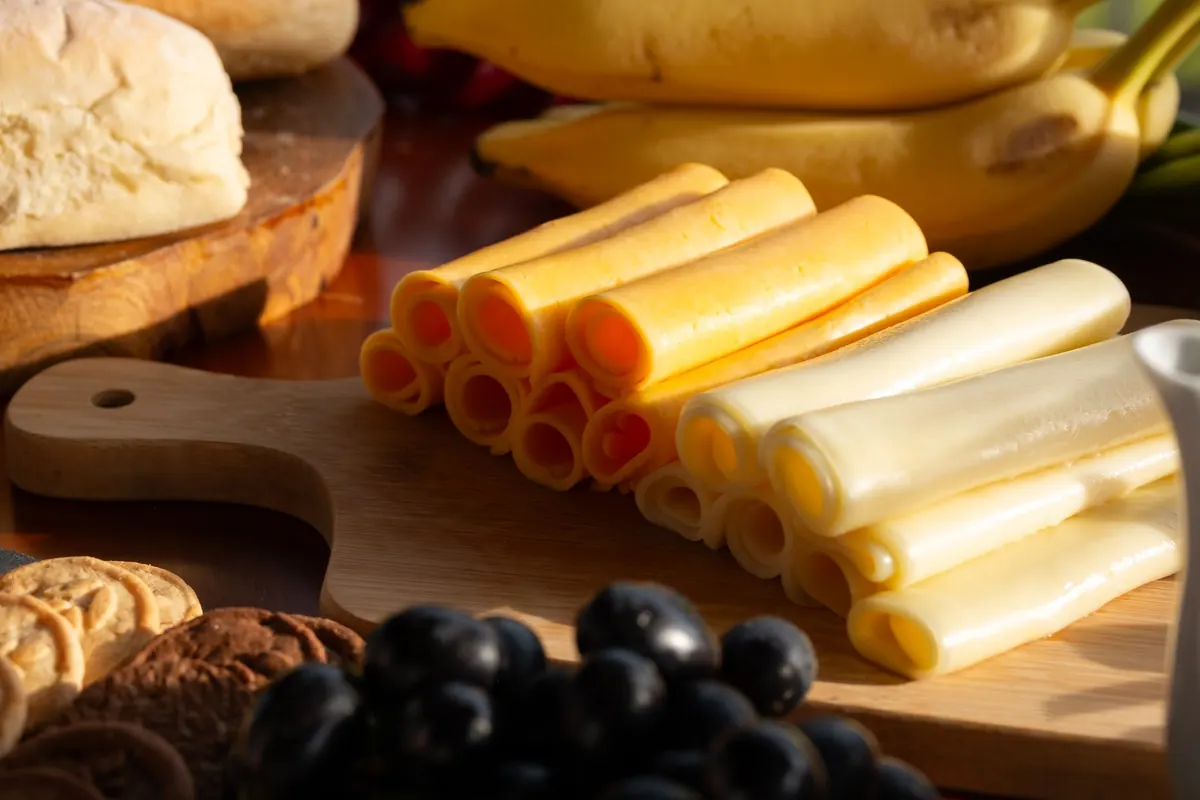 Tábua com queijos fatiados em rolinhos, pão, bananas, uvas e biscoitos
